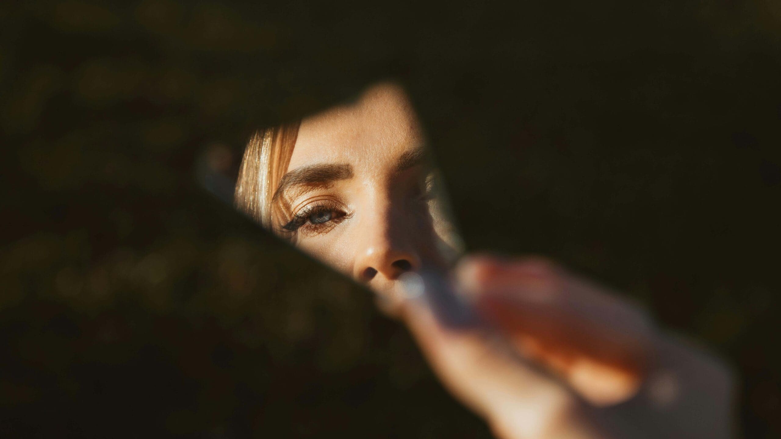 A woman's face reflects in a small mirror fragment against a dark background. Could trauma be quietly driving your codependent patterns? Online relational trauma therapy in Palm Beach, FL can help you find clarity.