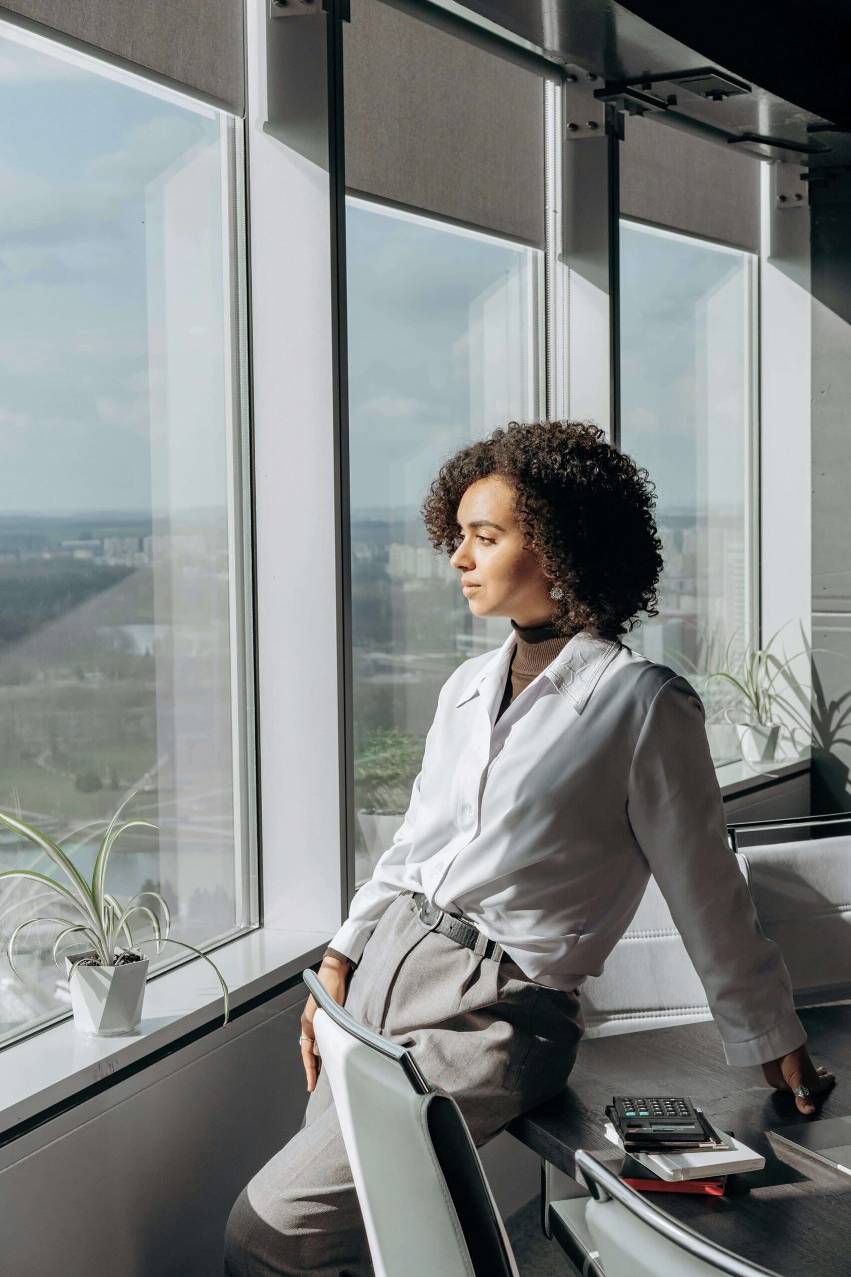 A professional woman leans against a desk near large windows. Is trauma from past relationships shaping your urge to constantly meet expectations? A relational trauma therapist in Palm Beach, FL, supports healing from these deeper emotional patterns.