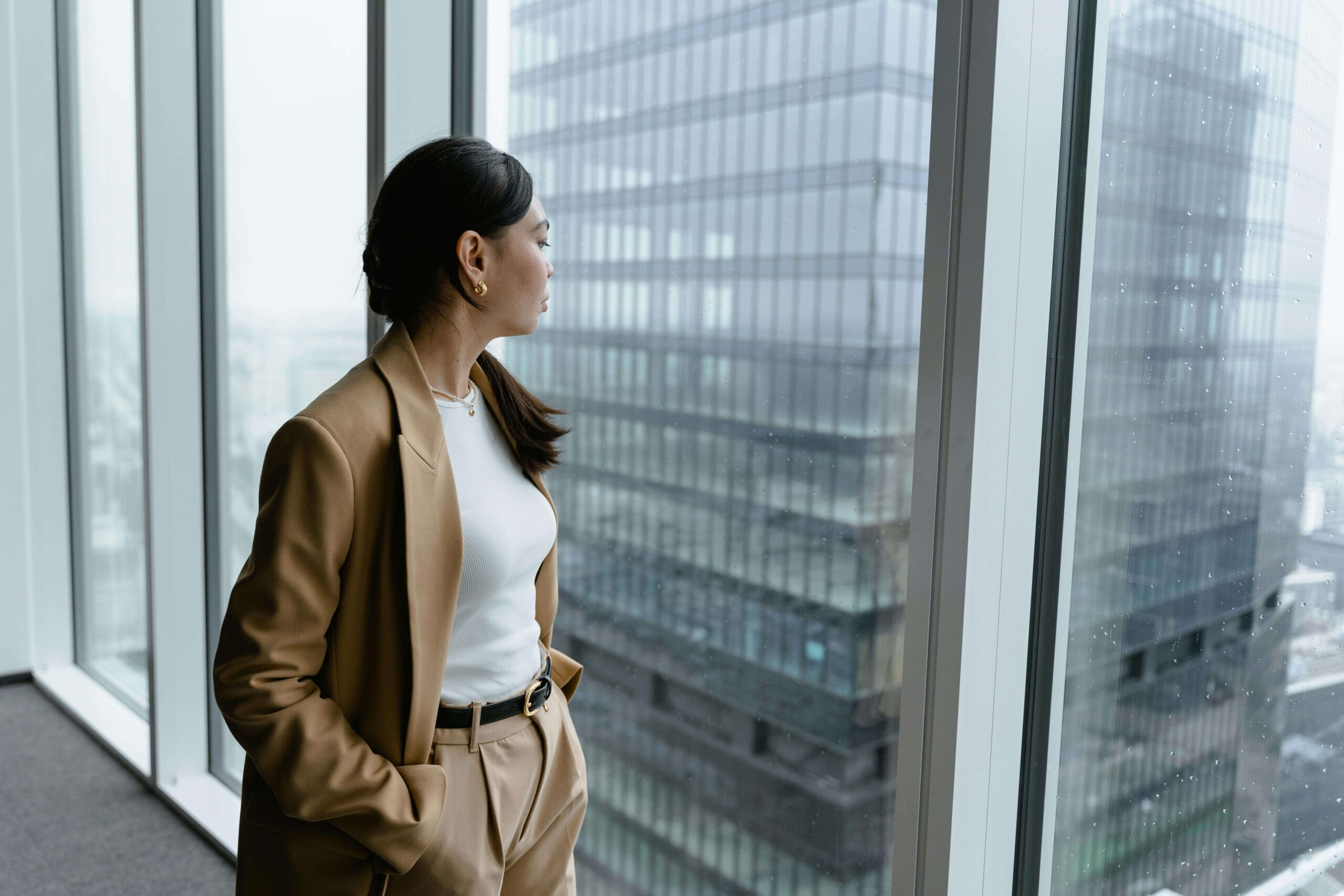 A professional woman gazes out of a high-rise office window. Could perfectionism be masking unresolved trauma that keeps you stuck in survival mode? A relational trauma therapist in Palm Beach, FL, can help you understand the deeper patterns driving your need to achieve.