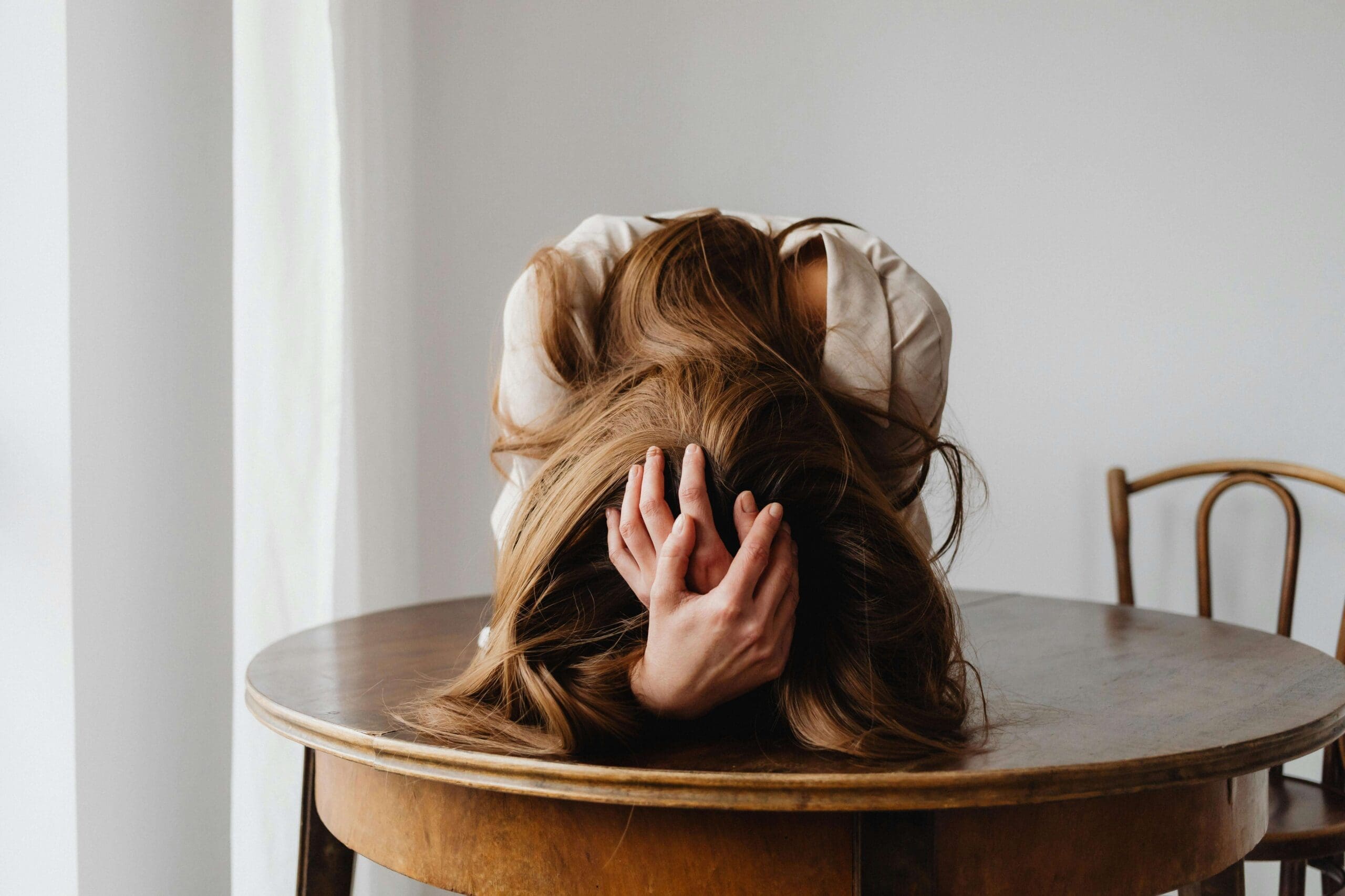A woman slumps forward onto a wooden table. Is unhealed trauma making parenting feel impossible? A relational trauma therapist in Palm Beach, FL can help you find relief, healing, and a more sustainable path forward for you and your family.
