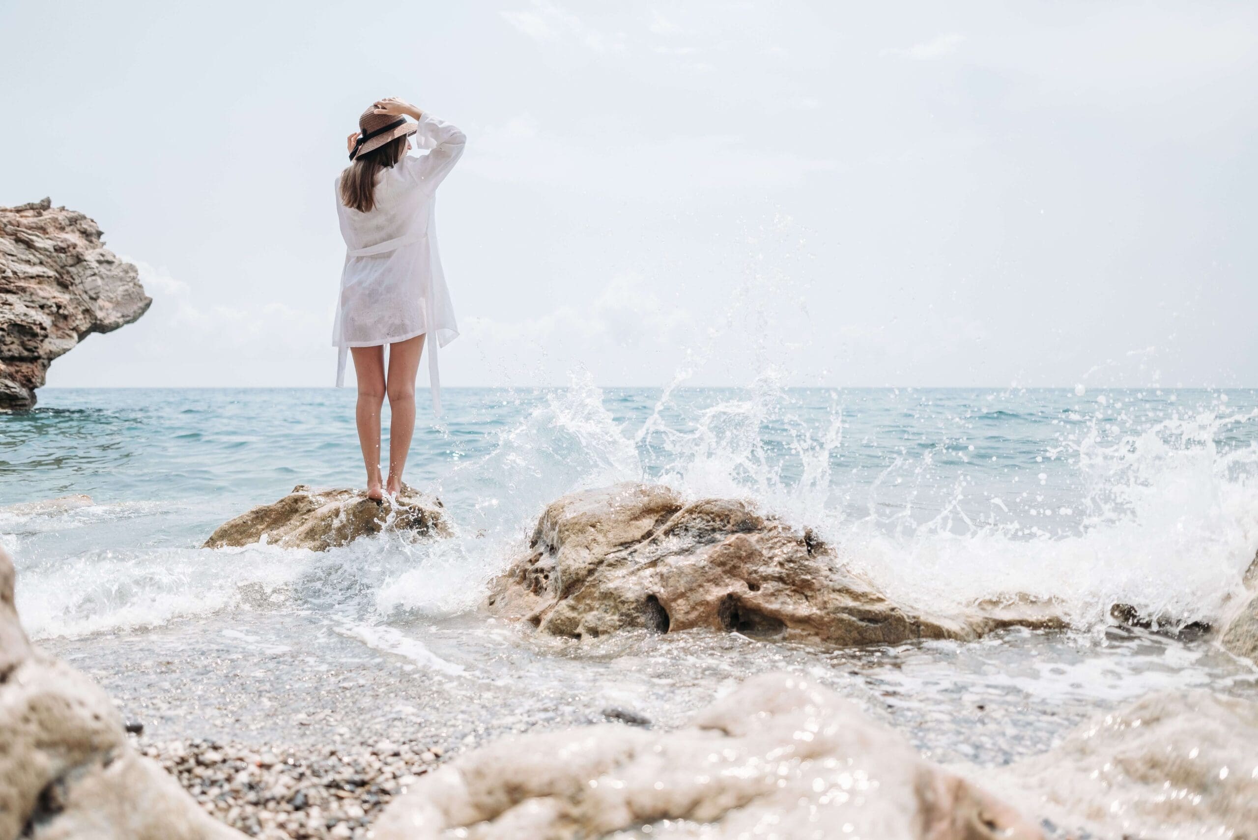 A woman in a straw hat stands on a coastal rock as waves crash. Are high-achieving women like you using the busyness of summer to avoid slowing down and facing what's underneath the surface? A relational trauma therapist in Palm Beach, FL, can help you find out.