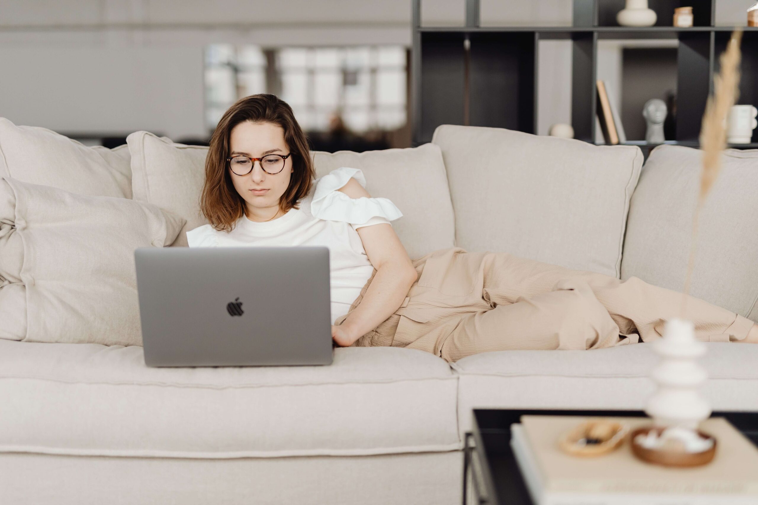 A woman in glasses works on her laptop while relaxing on a cozy couch. How can a therapist help when you feel stretched too thin? Online relational trauma therapy in Palm Beach, FL meets you right where you are.