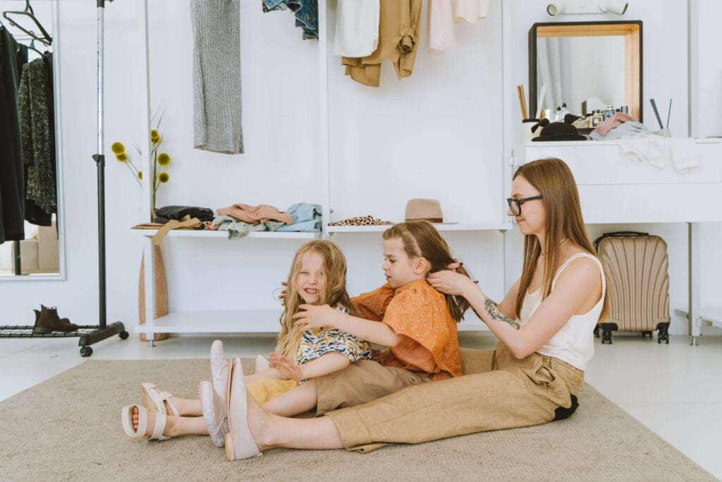 A mother sits on the floor braiding her daughters' hair as they get ready together. Does back-to-school season leave you stretched thin trying to do it all? Online relational trauma therapy in Palm Beach, FL helps you reconnect amid the busyness.