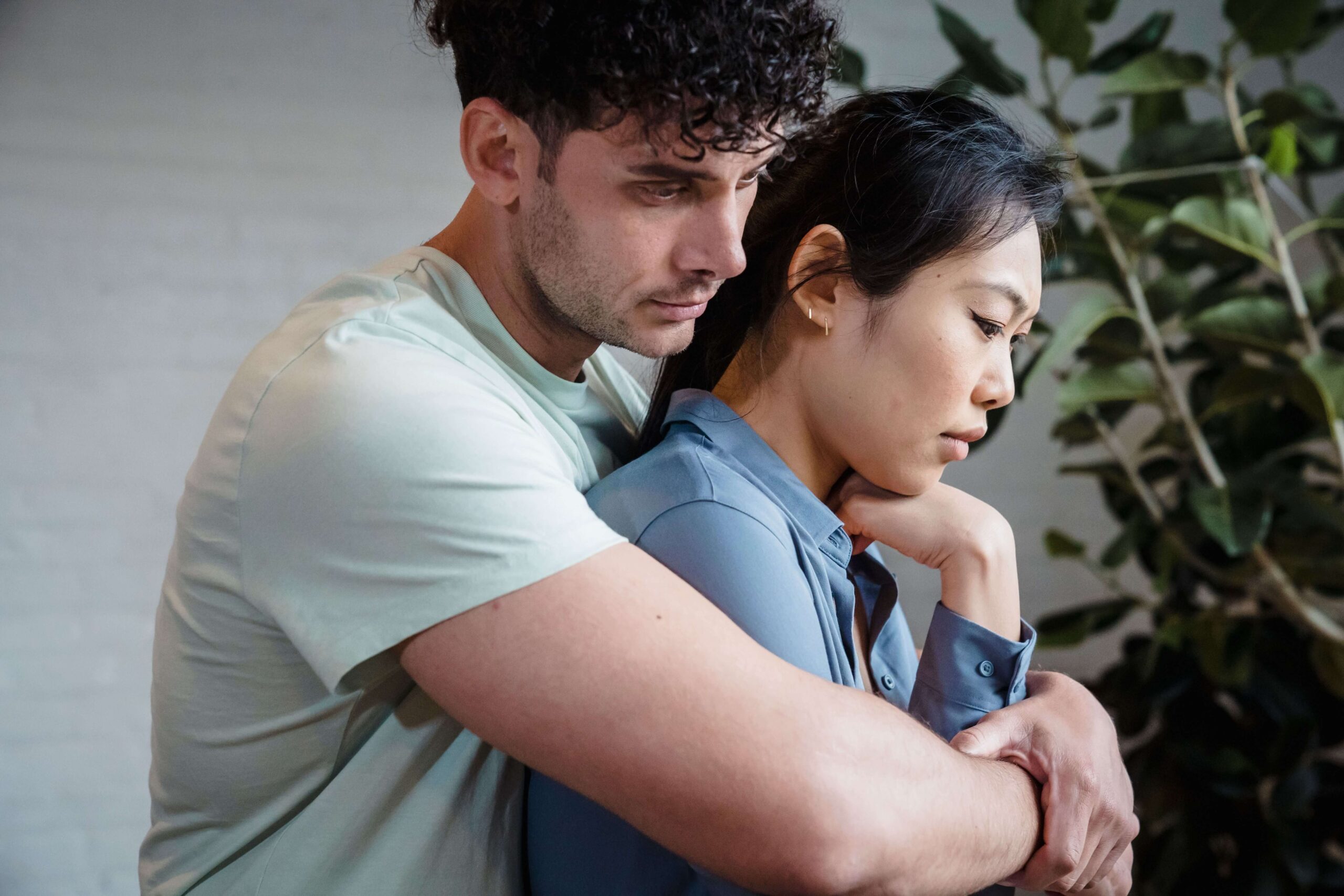 A man embraces a woman from behind while she gazes away with a distant expression. Does closeness sometimes feel more like a threat rather than a comfort in your relationships? An online relational trauma therapist in Palm Beach, FL can help you navigate emotional shutdown.